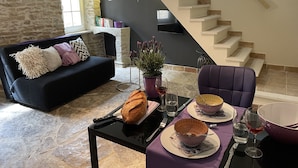 Un salon avec un mur en pierre, un canapé noir, une table à manger avec une miche de pain, des bols et des verres à vin.