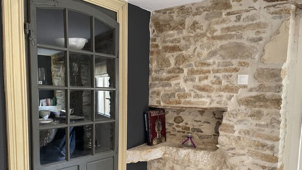 Une pièce avec un mur en pierre, une porte sombre dotée de panneaux de verre et une étagère sur laquelle se trouvent un livre et une petite bouteille.