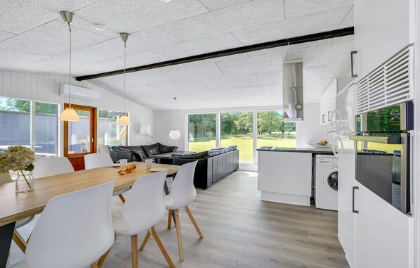 A modern kitchen with white cabinetry, a dining area with a wooden table and white chairs, and a view of a green garden through large windows.