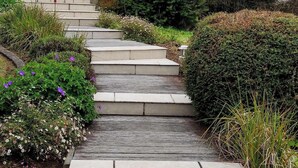 Een modern huis met twee verdiepingen, een beige gevel, donker dak en een balkon. Er zijn treden die leiden naar de ingang, omringd door groen en bloemen.