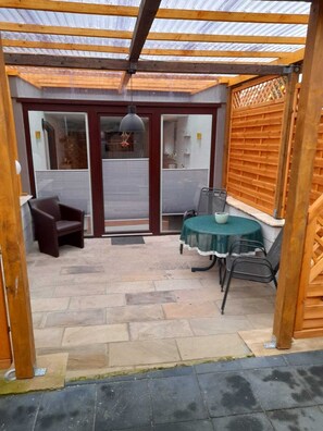 A covered patio area with a table and chairs, a door leading to a room, and a wooden fence. - Ferienwohnung Familie Hohm (Lohmen)