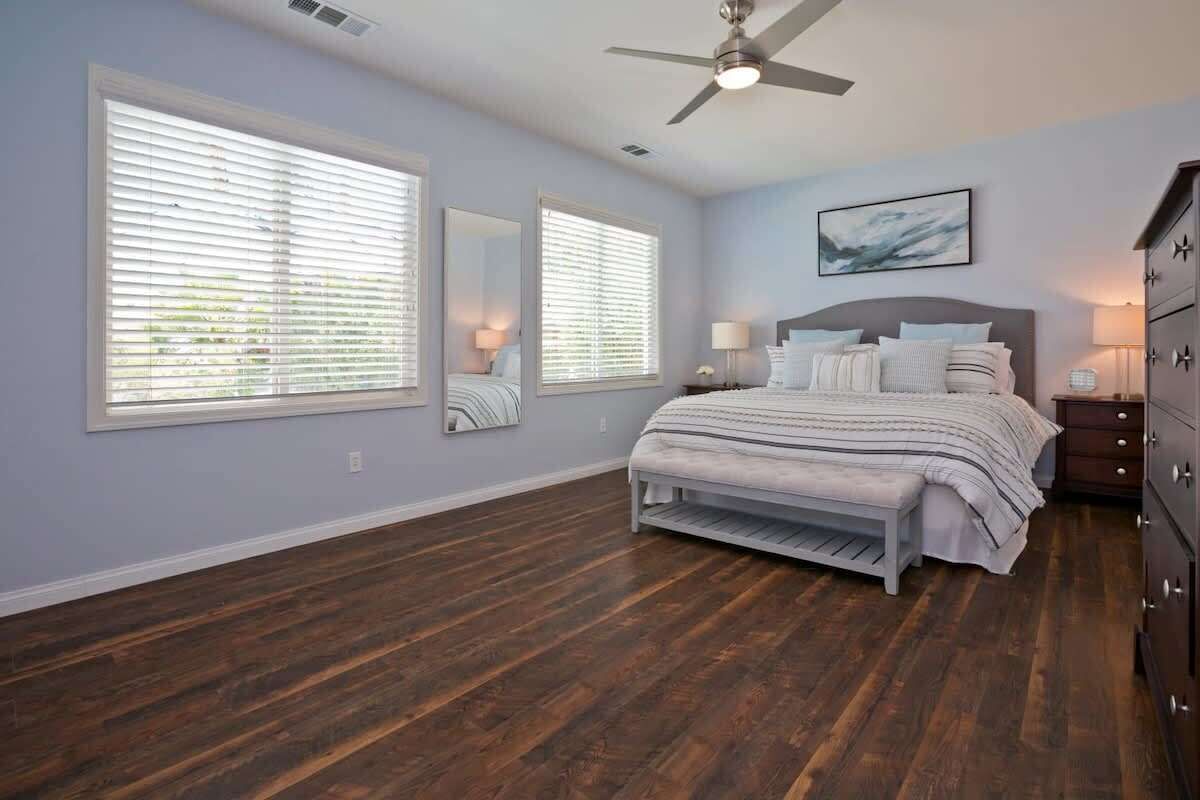 A bedroom with a bed, two bedside tables, a dresser, a ceiling fan, and large windows with blinds.