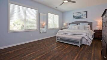 A bedroom with a bed, two bedside tables, a dresser, a ceiling fan, and large windows with blinds.