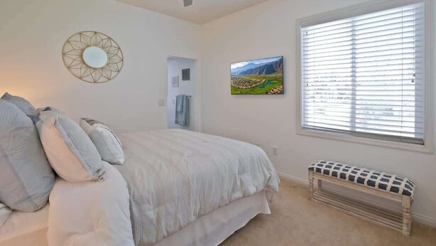 Une chambre à coucher comprenant un lit, un ventilateur de plafond, une fenêtre avec des stores, un banc et un miroir fixé au mur.