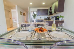 Un comedor con una mesa de vidrio preparada para dos personas, una botella de vino y una bandeja de fruta.