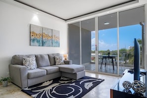 A modern living room with a grey sectional sofa, a large glass sliding door leading to a balcony, and a piece of abstract art on the wall. - 2BR Condo w/ Balcony & Pool near Eagle Beach (Eagle Beach)