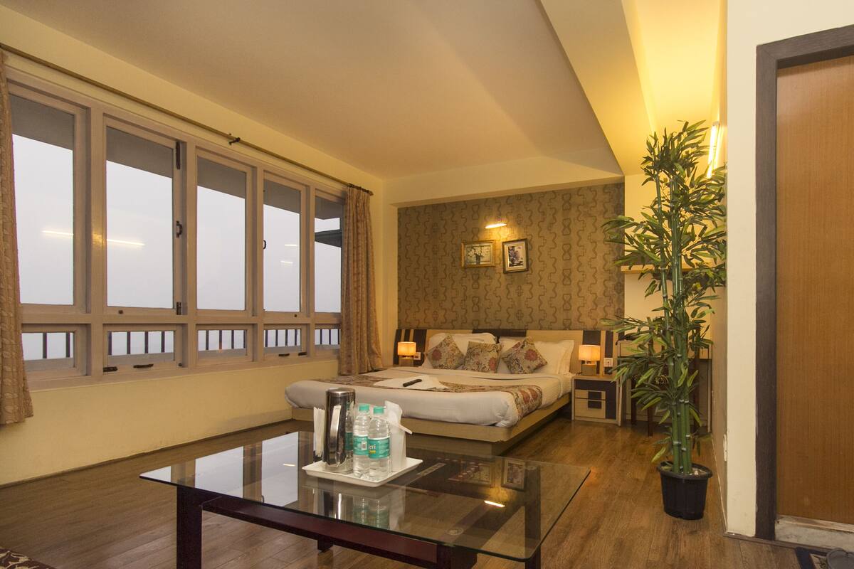 Une chambre d’hôtel avec une grande fenêtre, un lit recouvert d’une literie à motifs floraux, une table de chevet avec une lampe et une plante en pot.
