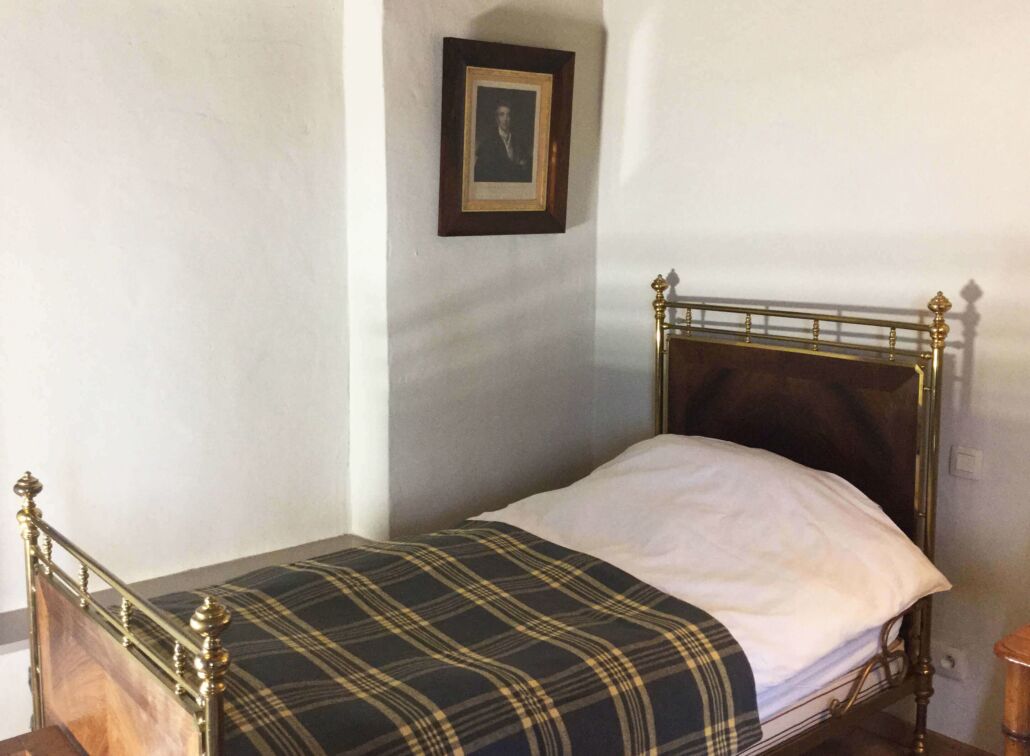 A single bed with a plaid bedspread and a white pillow in a room with plain walls and a framed picture on the wall.