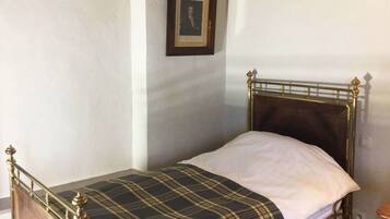 A single bed with a plaid bedspread and a white pillow in a room with plain walls and a framed picture on the wall.