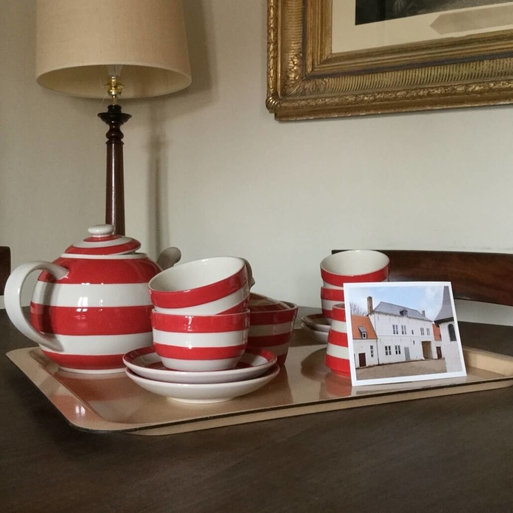 Une table mise avec une théière, des tasses et des soucoupes à rayures rouges et blanches, une lampe et un tableau représentant une maison.