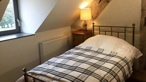 Un dormitorio con techo inclinado, cama con colcha escocesa, lámpara de noche y ventana con vistas a árboles.