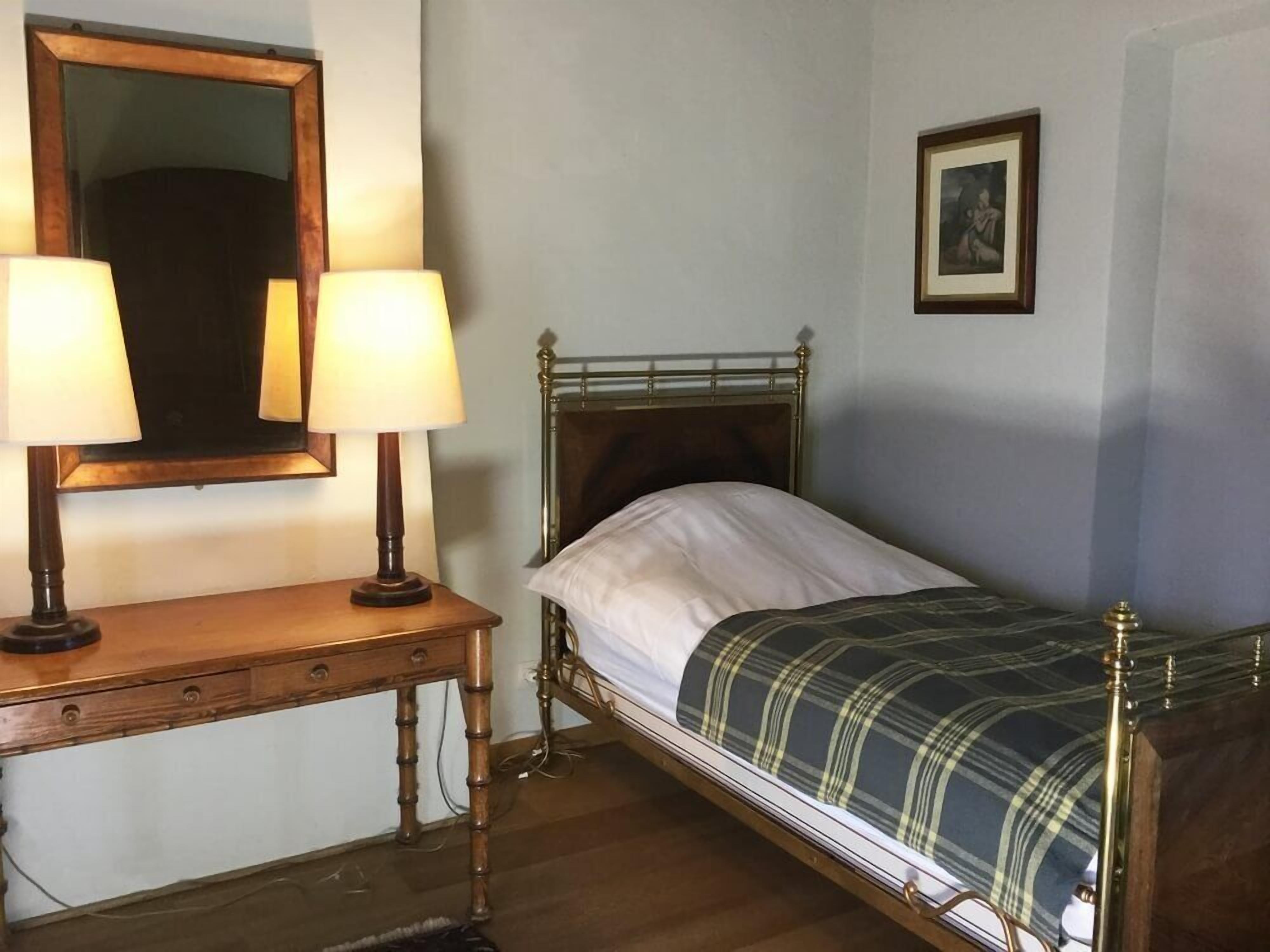 A single bed with a metal frame and a plaid blanket, a wooden bedside table with two lamps, a mirror, and a framed picture on the wall.
