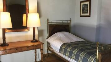 A single bed with a metal frame and a plaid blanket, a wooden bedside table with two lamps, a mirror, and a framed picture on the wall.