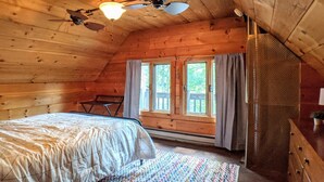 A cozy attic bedroom with a bed, a colorful rug, a ceiling fan, and two windows with curtains. - Lakes End Log Cabin on Lake Wicwas! (Meredith)