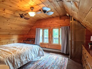A cozy attic bedroom with a bed, a colorful rug, a ceiling fan, and two windows with curtains.