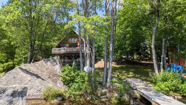 A cabin with a deck, surrounded by trees and a small stream. - Lakes End Log Cabin on Lake Wicwas! (Meredith)