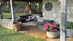 An outdoor patio with a stone wall, a wooden pergola, a wicker sofa, a table, and a large potted plant with pink flowers.