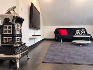 A living room with a vintage stove, a flat-screen TV, a sofa with red and patterned cushions, and a small table with a tray.