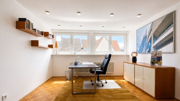 A modern office space with a desk, chair, shelving, radiator, and a large window with a view of buildings.