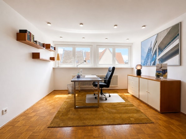 A modern office space with a desk, chair, shelving, radiator, and a large window with a view of buildings.