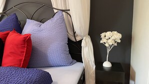 A bed with a metal frame, white and purple pillows, and a white canopy. There is a small black nightstand with a white lamp and a white flower arrangement. - Paradis de Provence (Le Pin)