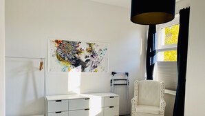 Une chambre avec une commode blanche, une chaise, un radiateur et une fenêtre avec des stores.
