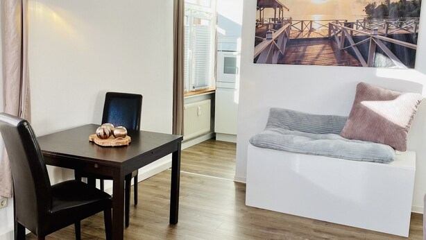 Un salon moderne avec un coin repas, comprenant un plancher en bois, un canapé blanc avec des coussins, une table noire agrémentée d’un élément décoratif central, et un grand tableau représentant un coucher de soleil sur une jetée.