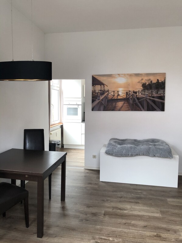 A modern dining area with a wooden table and chairs, a white bench with a gray blanket, and a sunset painting on the wall.