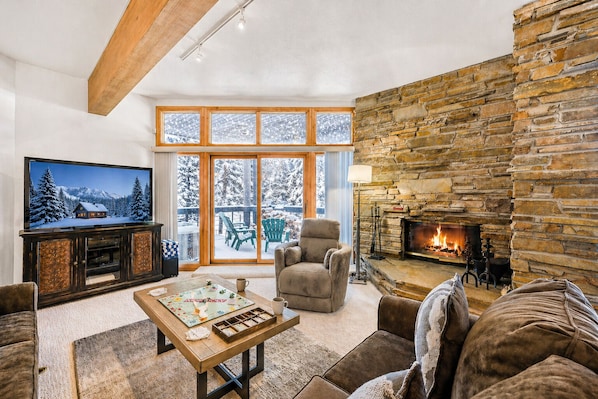 A cozy living room with a stone fireplace, a large TV, and a view of snow-covered trees through the windows. - 12 C Powder Run 2 Bedroom Condo by RedAwning (Park City)