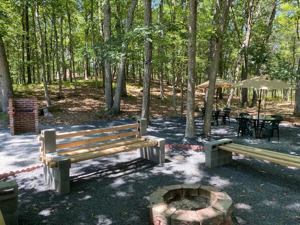 A forested area with a wooden bench, picnic tables, and a fire pit.