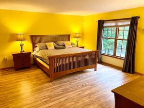 A bedroom with a wooden bed, two bedside tables, a window with curtains, and a view of greenery outside.