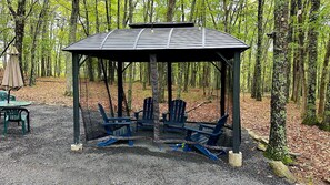 A gazebo with a roof, surrounded by chairs, set in a forested area with trees and gravel ground. - Bushkill Beauty-HotTub, Firepit, Lake, Canoe, Pool (Lehman township)