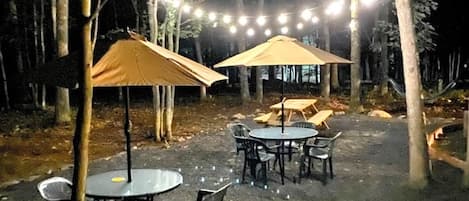Un área de comedor al aire libre con mesas y sillas, rodeada de árboles, y luces colgantes.