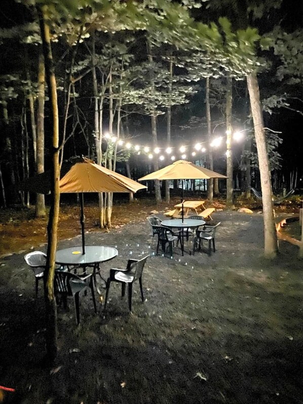 An outdoor dining area with tables and chairs, surrounded by trees, and string lights overhead.