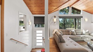 A modern living room with a wooden ceiling, a beige sectional sofa, and large windows offering a view of trees. - Sunset Villa: Upscale Three Bedroom Three Bath with Ocean Views (Pebble Beach)