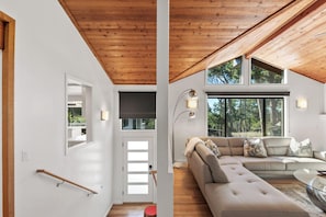 Una sala de estar moderna con techo de madera, un sofá seccional beige y grandes ventanas que ofrecen vista a árboles.