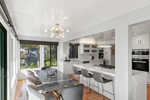 A modern kitchen with white cabinets, a central island with bar stools, and a large window providing a view of the outdoors. - Sunset Villa: Upscale Three Bedroom Three Bath with Ocean Views (Pebble Beach)