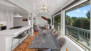A modern kitchen with a marble countertop, a dining area with a large table, and a view of the outdoors through large windows. - Sunset Villa: Upscale Three Bedroom Three Bath with Ocean Views (Pebble Beach)