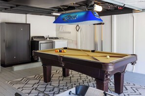 A pool table with a blue overhead light, a washing machine, and a cabinet in a room with a patterned rug. - Pacific Panorama: Stunning Ocean Views and Pacific Sunsets (Carmel)