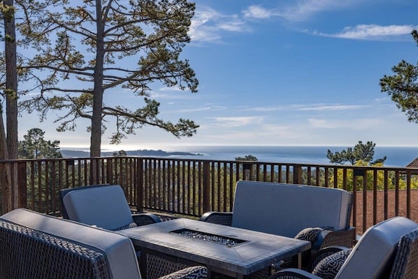 A patio with wicker furniture and a fire pit, overlooking a scenic view of the ocean and hills.