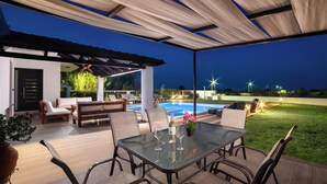 An outdoor patio with a glass table, chairs, and a pool in the background. - Elysian Villa in Afantou (Αφάντου)