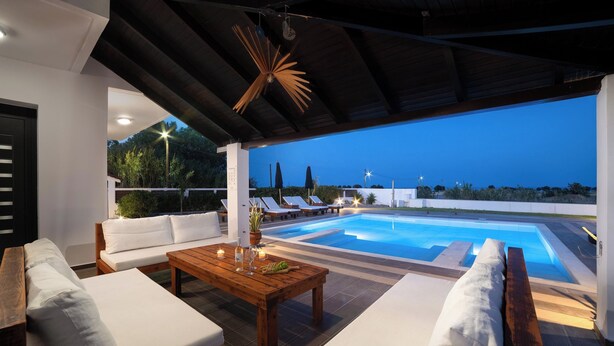 A poolside area with a wooden table, white cushioned seating, and a view of the pool at dusk. - Elysian Villa in Afantou (Αφάντου)
