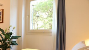 Une chambre d’hôtel avec un lit, une fenêtre avec des rideaux, une plante en pot et un luminaire fixé au mur.