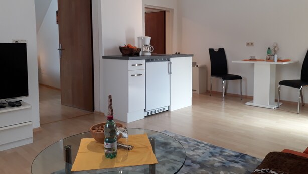 A modern living room with a glass coffee table, a TV, and a dining area with a white table and two chairs.