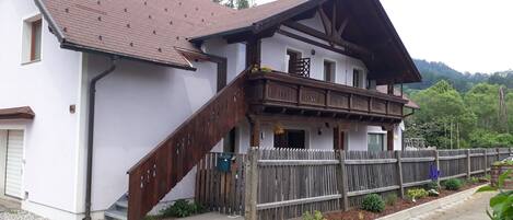 Un edificio de dos pisos con escalera y balcón de madera, rodeado por una valla de madera y macizos de flores.