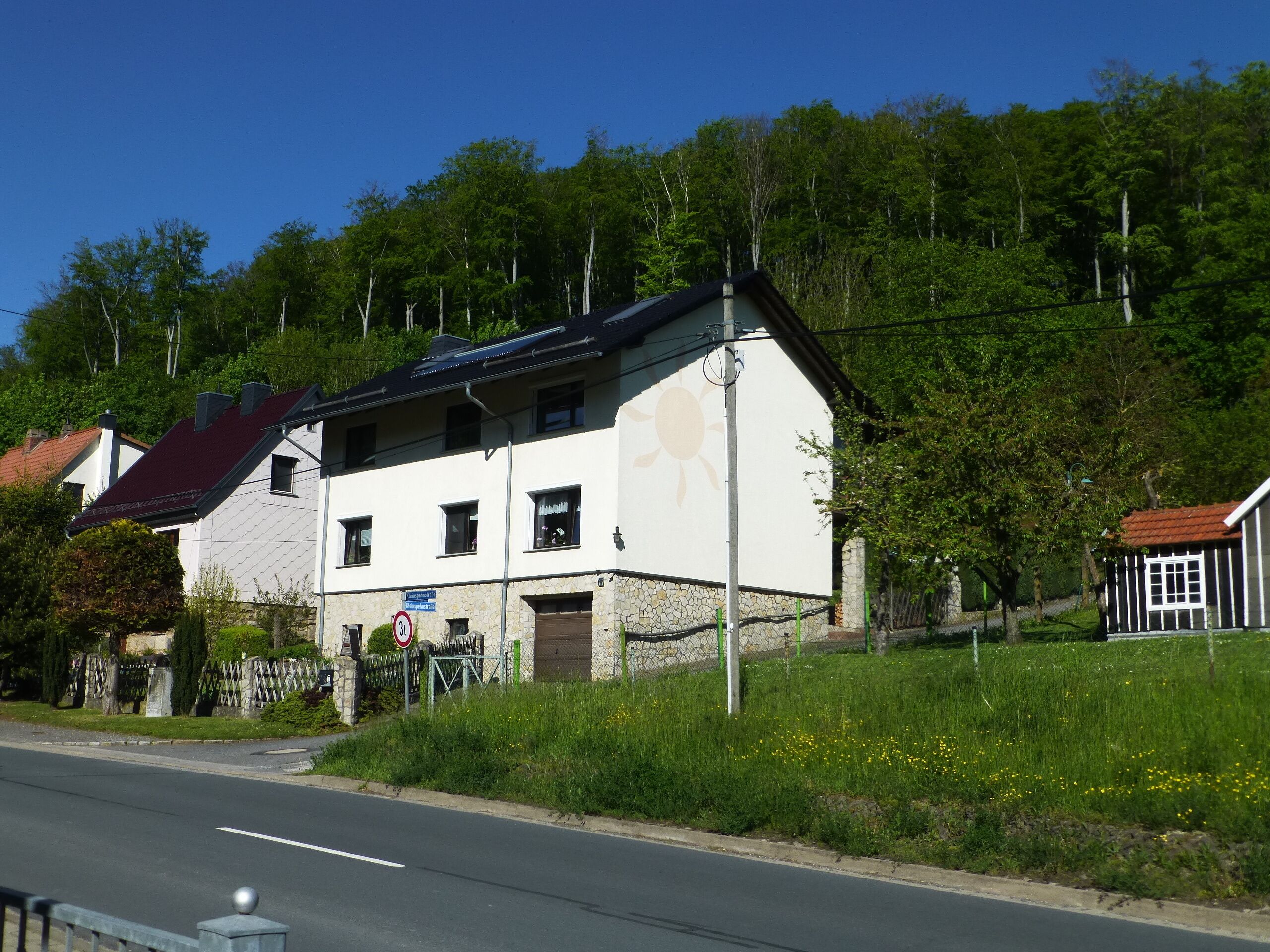 Ein zweistöckiges Gebäude mit weißer Fassade und rotem Dach, neben einer Straße, über der ein klarer, blauer Himmel zu sehen ist.
