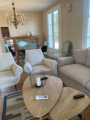 A living room with a sofa, armchairs, a coffee table, a chandelier, a dining table with chairs, and a view through a window.