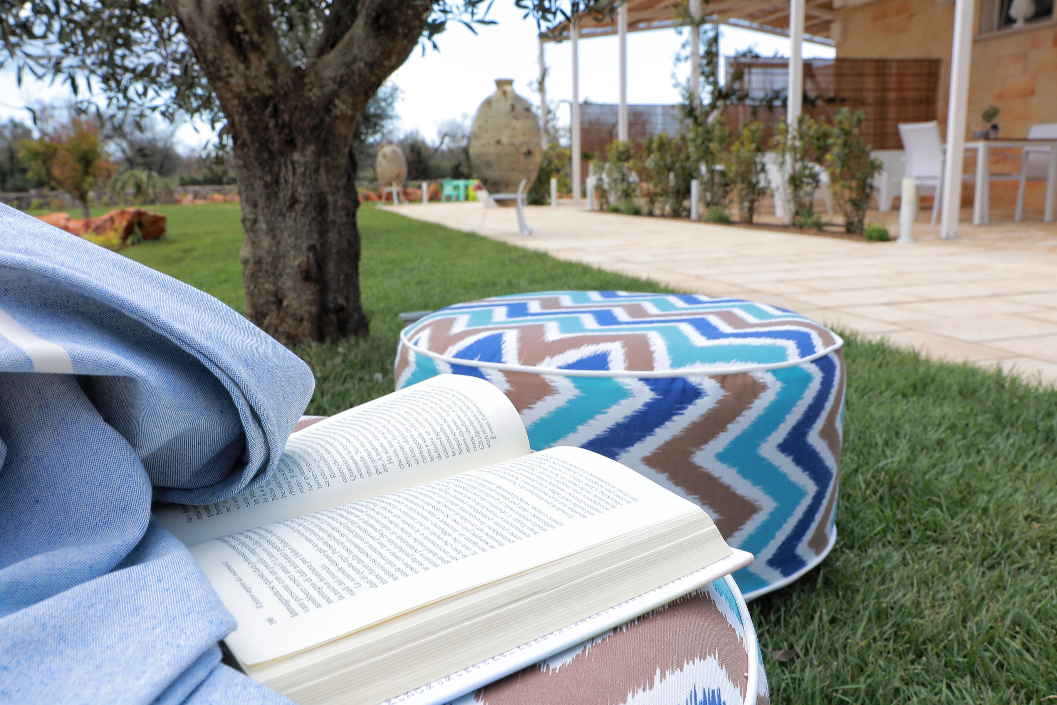 A book on a patterned cushion on grass, with a tree and outdoor furniture in the background.