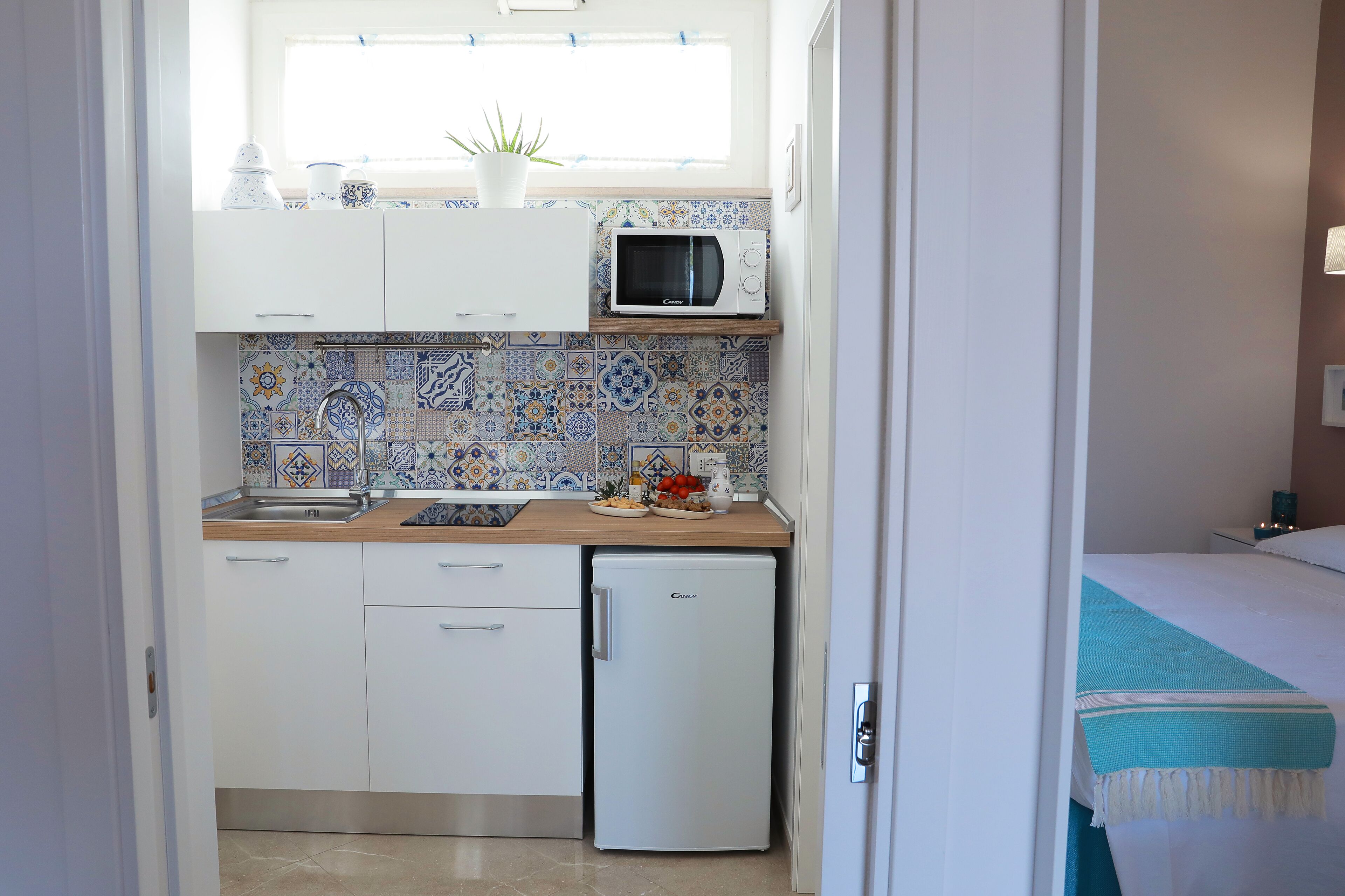 Una cocina compacta con microondas, fregadero y un refrigerador pequeño. Hay una ventana sobre el fregadero y una planta en maceta en la encimera.
