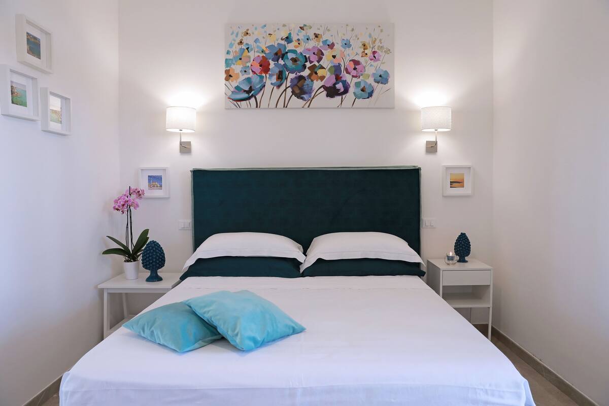 Ein ordentlich bezogenes Bett mit weißen und türkisfarbenen Kissen, ein Blumenbild darüber und zwei Nachttische mit Lampen.